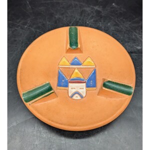 May include: Round, orange ceramic ashtray with a central design featuring a stylized face and geometric shapes in blue, yellow, and white. Green rectangular accents are placed around the rim. A vintage decorative piece.
