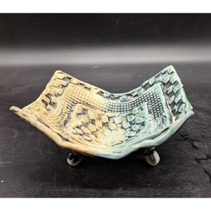 May include: A small, square ceramic dish with a geometric pattern in shades of green, brown, and white. The dish has three small feet and is perfect for holding jewelry or other small items.