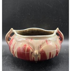 May include: A decorative ceramic bowl with a cream and red glaze. The bowl has two handles and a unique shape. The red glaze drips down the sides of the bowl, creating a striking visual effect. The bowl is likely vintage or antique.