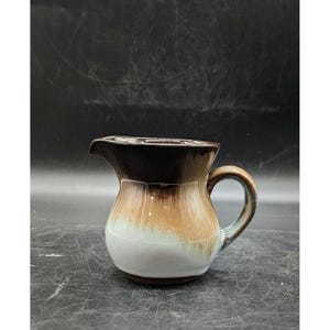 May include: A brown and white ceramic creamer with a glazed finish. The creamer has a rounded body and a handle.