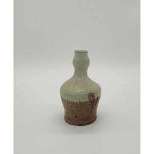 May include: A handmade ceramic vase with a bulbous base and a slender neck. The vase features a two-tone design, with a light green upper portion and a textured brown lower section. A brown feather design is painted on the side.