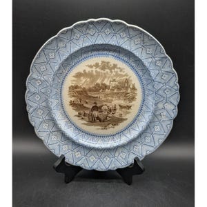 May include: A round ceramic plate with a scalloped edge featuring a light blue and white lattice pattern on the rim. The center displays a sepia-toned illustration of a riverside landscape with a person, boats, and buildings.