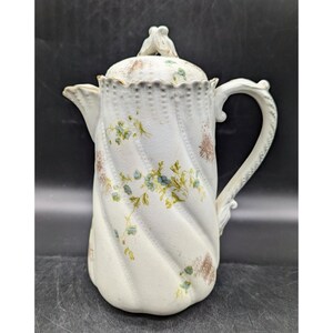 May include: White porcelain coffee pot with a floral design. The pot has a lid with a decorative knob and a twisted body. The handle is ornate and has a floral design.