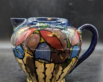 May include: A small, blue and white ceramic creamer with a floral design. The creamer has a handle and a spout. The floral design is in shades of red, blue, and purple.