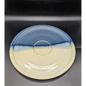 May include: A handmade ceramic plate with a blue and cream color scheme. The plate features a circular indentation in the center and a speckled texture. The top half is blue, transitioning to cream on the bottom.