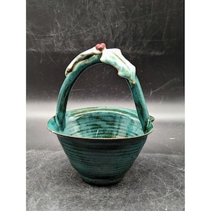 May include: A handcrafted teal ceramic basket with a handle. The basket has a textured surface and a glossy finish. The handle is decorated with a leaf and berry design. The basket is suitable for decorative use.