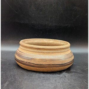 May include: A handcrafted, round ceramic bowl with a wide, open top. The bowl features a textured, wave-like pattern in shades of brown and tan around the exterior. The interior and rim are a lighter tan color. The bowl sits on a dark surface.