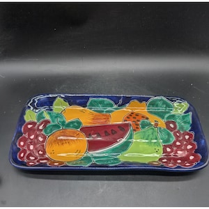 May include: A rectangular ceramic serving tray featuring a hand-painted design of assorted fruits including a slice of watermelon, an orange, a pear, and clusters of grapes, all set against a deep blue background with green leaves.