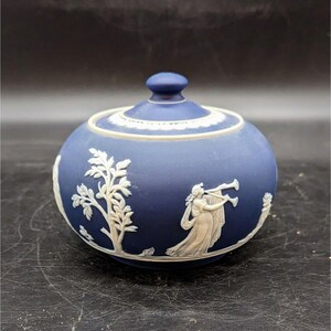 May include: A blue and white ceramic sugar bowl with a lid. The bowl is decorated with a white relief design of a woman playing a horn and a tree.