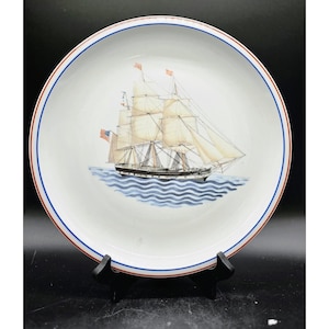 May include: White ceramic plate with a detailed illustration of a tall ship sailing on blue waves. The plate features a red and blue border. The ship has multiple sails and an American flag. A decorative nautical item.