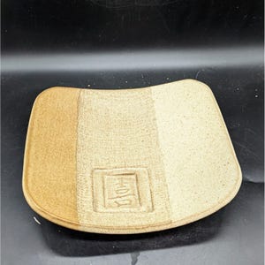 May include: A square ceramic serving dish with a light brown, tan, and white glaze. The dish has a slightly curved edge and a raised rectangular area in the center with a Chinese character.