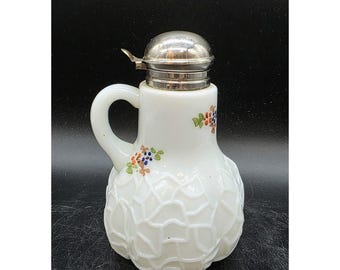 Può includere: Lattiera in vetro bianco con coperchio e manico argentati. La lattiera ha un motivo strutturato a forma di ragnatela sul fondo ed è decorata con piccoli grappoli floreali nei toni del blu, arancione e verde. Un pezzo classico per servire panna o latte.