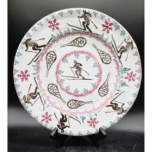 May include: A white ceramic plate with a winter sports theme. The plate features skiers, snowshoes, and snowflakes in brown, pink, and blue. The rim is pink, and the plate is displayed on a black stand.