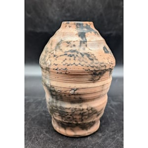 May include: A handmade, terracotta-colored vase with a unique, layered design. The vase features a cylindrical shape with a slightly flared top and base. Dark, mottled patterns add visual interest to the textured surface.