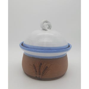 May include: A ceramic lidded jar with a brown base and a white lid. The base features a wheat stalk design. The lid has a blue stripe and a swirl handle. The jar is likely handmade and suitable for kitchen storage.
