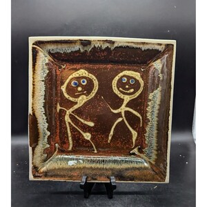 May include: Square ceramic plate with a brown glaze and a white rim. Two stick figures with blue eyes are drawn on the plate.