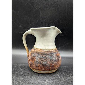 May include: A handmade ceramic jug with a cream-coloured top and a brown, textured body. The body features a wave-like pattern. The jug has a curved handle and a spout for pouring. A rustic, decorative piece.