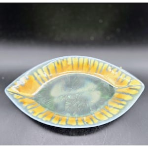 May include: An oval-shaped ceramic dish with a light blue rim and a central design in shades of green and yellow. The dish has a textured surface and is likely used for serving or display.
