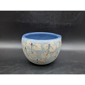 May include: A round, light blue ceramic planter with a dark blue interior. The exterior features a repeating pattern of white leaves with black stems. The planter is suitable for small plants and succulents.