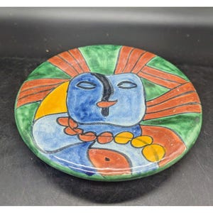 May include: A round ceramic plate with a colourful abstract design. The design features a blue face with closed eyes, a red mouth, and a yellow and orange background. The plate is decorated with red and orange stripes.