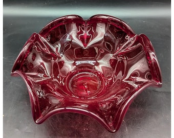 May include: A ruby red glass bowl with a unique, scalloped edge design. The bowl has a central, circular indentation and a series of decorative, pointed edges. The glass is transparent, allowing light to pass through, and is likely vintage.