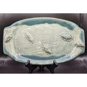 May include: A handmade ceramic serving platter with a light blue and teal glaze. The oval-shaped platter features raised leaf-like designs and a decorative line pattern. The edges are slightly raised, and the platter is displayed on a black stand.