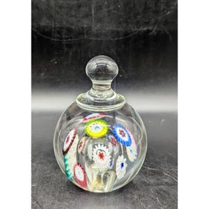 May include: A clear glass decorative bottle with a round body and a bulbous stopper. The bottle is filled with colorful floral designs in red, white, blue, and yellow. The bottle is likely used for perfume or decorative purposes.