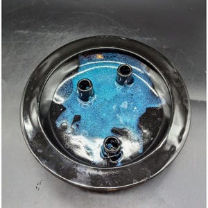 May include: A round, black ceramic candle holder with a glossy finish. The interior features a vibrant blue and turquoise glaze with three black candle holders. The candle holder is designed to hold three candles.