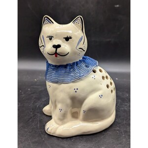 May include: A ceramic cat figurine in a sitting position. The cat is off-white with blue accents, including a blue collar and small blue dots. The cat has a smiling face with a red tongue and several holes on its side.
