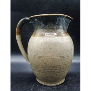 May include: A handcrafted stoneware jug with a wide base and a curved handle. The jug features a speckled, light beige body with a dark brown rim and handle. Horizontal lines encircle the body, adding texture to the rustic design.