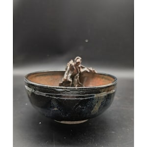May include: A round ceramic bowl with a dark, glossy black exterior and a textured, reddish-brown interior. A small, metallic-toned sculpted figure is attached to the inner rim, appearing to lean over the edge of the bowl into the center.