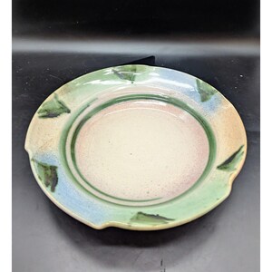May include: A handmade ceramic plate with a unique, organic design. The plate features a cream-colored center, a pink inner ring, and a green and blue outer rim. The rim has a textured, irregular shape with small, dark green accents. This decorative plate is perfect for serving food.