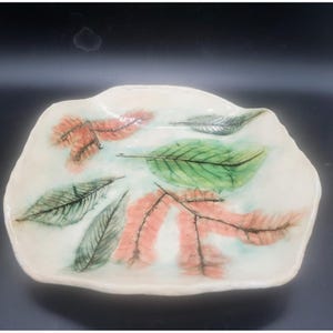 May include: A handmade ceramic serving dish with a white base. The dish is decorated with painted leaves in shades of green, orange, and gray. The dish has an irregular shape and is suitable for serving food or as a decorative piece.