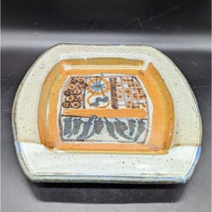 May include: A square ceramic serving dish with a raised rim and a speckled glaze. The dish features a hand-painted design of geometric shapes and stylized peppers in shades of brown, orange, and blue.