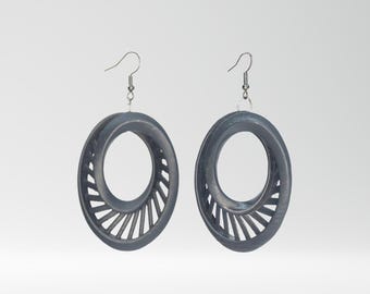 Può includere: Un paio di orecchini ovali grigio scuro. Ogni orecchino presenta un centro cavo con un design a ventaglio su un lato. Gli orecchini pendono da ganci per orecchie color argento.