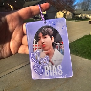 May include: A purple plastic keychain with a photo of a young man with dark hair. The keychain has three white hearts and the word "BIAS" printed on it.