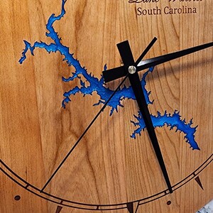 May include: A wooden clock with a blue outline of Lake Wateree, South Carolina. The clock has black hands and a compass rose with north, south, east, and west markings.
