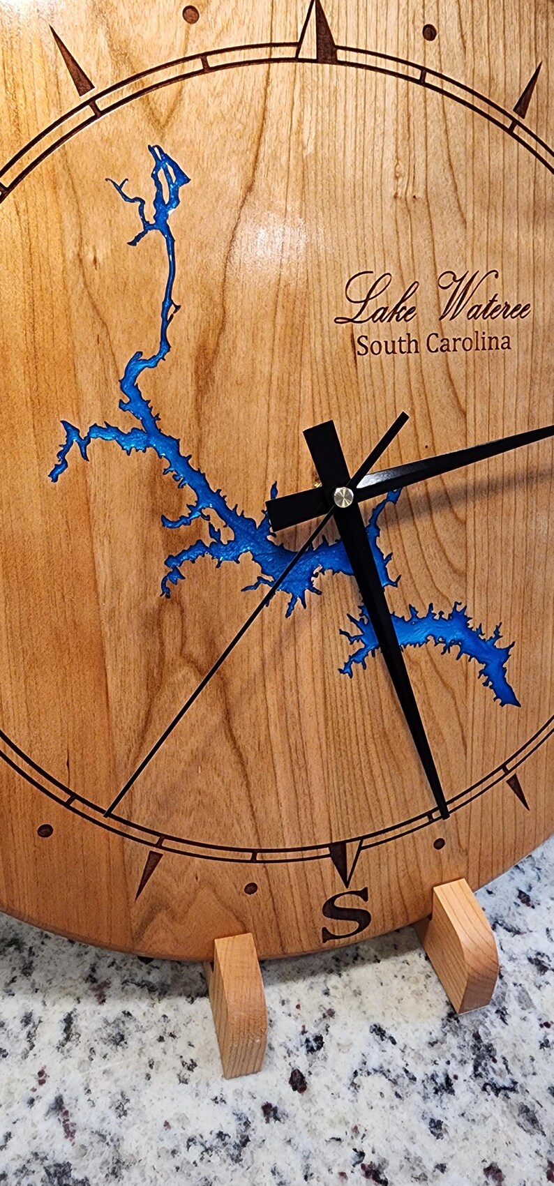 May include: A wooden clock with a blue outline of Lake Wateree, South Carolina. The clock has black hands and a compass rose with north, south, east, and west markings.