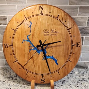 May include: A wooden clock with a compass design and a blue map of Lake Wateree, South Carolina. The clock has black hands and a gold center.