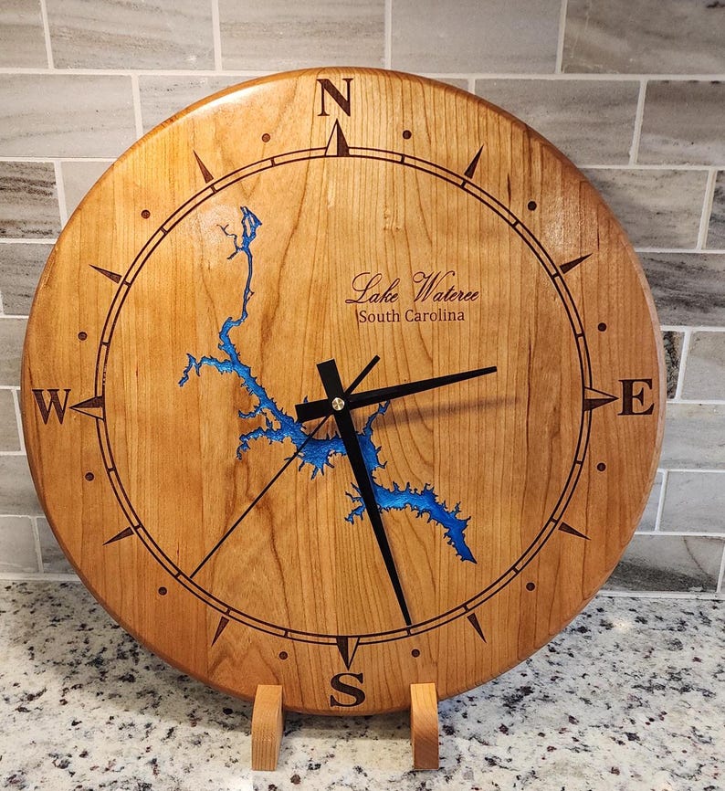 May include: A wooden clock with a compass design and a blue map of Lake Wateree, South Carolina. The clock has black hands and a gold center.