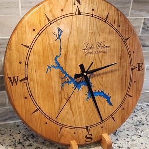 May include: A round wooden clock with a compass design and black hands. The clock face features a blue lake map and the text "Lake Wateree South Carolina". The clock is supported by a wooden stand.