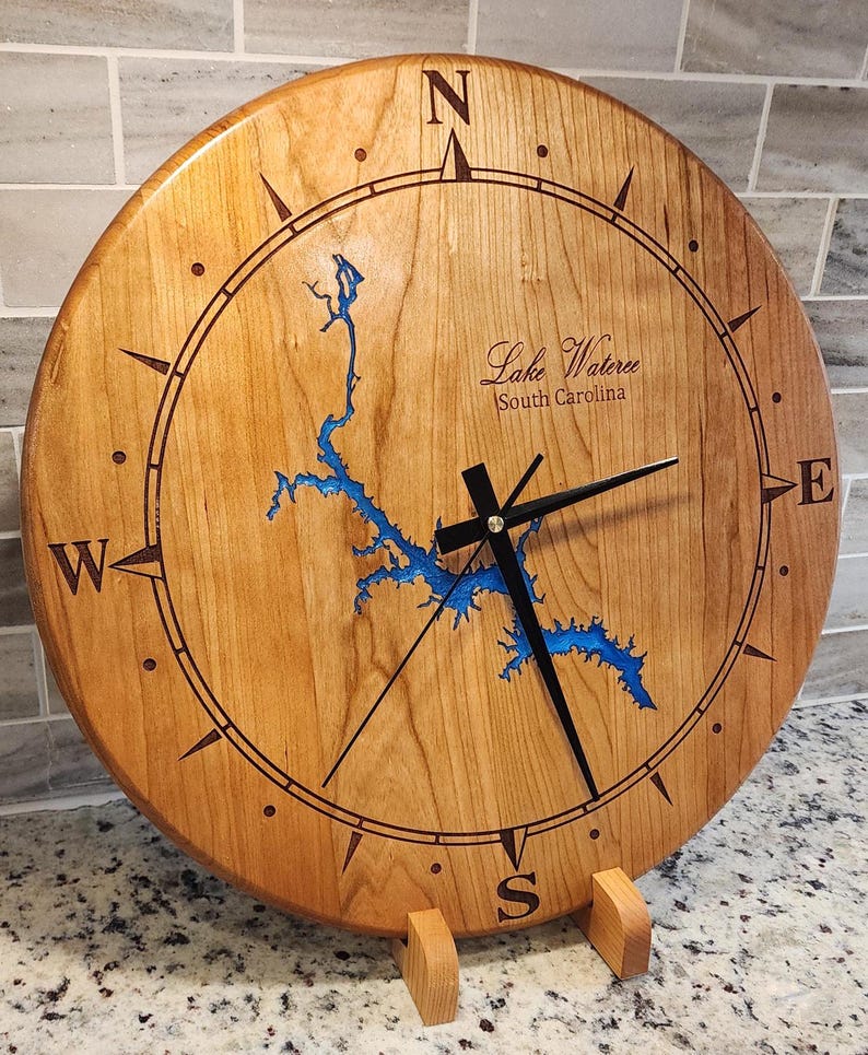 May include: A round wooden clock with a compass design and black hands. The clock face features a blue lake map and the text "Lake Wateree South Carolina". The clock is supported by a wooden stand.