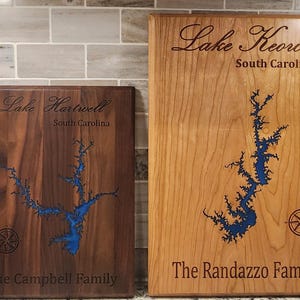 May include: Two wooden plaques with engraved lake outlines and family names. The plaque on the left is dark brown with the text "Lake Hartwell, South Carolina, The Campbell Family". The plaque on the right is light brown with the text "Lake Keowee, South Carolina, The Randazzo Family".