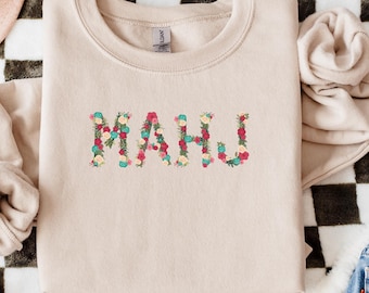 May include: A beige sweatshirt with the word "MAHJ" embroidered in colorful floral letters. The sweatshirt is laid flat on a black and white checkered surface, with a pink hat and a pair of blue jeans in the background.
