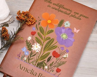 Puede incluir: Un diario marrón con un diseño floral con flores naranjas, moradas y rojas, y el texto "Como las flores silvestres, crecemos en lugares inesperados". El nombre "Amelia Rose" está impreso en la parte inferior.
