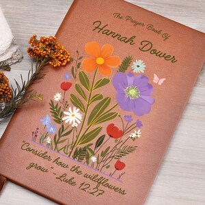 Könnte beinhalten: Ein braunes, ledergebundenes Tagebuch mit einem floralen Design und dem Text "The Prayer Book Of Hannah Dower" und "Consider how the wildflowers grow - Luke 12:27".