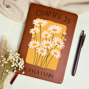 May include: A brown leather-bound journal with the text "CHAPTER 35" and "KAYLA LYNN" printed on the cover. The cover features a yellow rectangle with a daisy floral design. A dark pen rests on the right side of the journal.