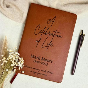 May include: A brown leather journal with the text "A Celebration of Life" and the name "Mark Moser" with dates 1949-2025. A pen and dried flowers are next to the journal. The journal also has the text "Share a memory, words of love or sign your name."