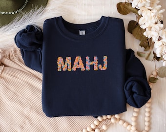 May include: Navy blue sweatshirt with the word "MAHJ" embroidered in colorful floral letters. The sweatshirt has long sleeves and a crew neck. The background includes a hat, flowers, and decorative beads.
