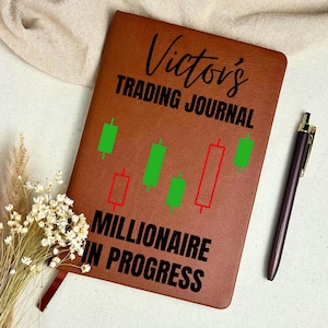 Könnte beinhalten: Ein braunes, ledergebundenes Notizbuch mit dem Text "Victor's Trading Journal" und "Millionaire in Progress" in Schwarz. Das Notizbuch zeigt grüne und rote Börsenkerzen-Grafiken. Ein dunkler Stift liegt neben dem Notizbuch.